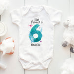 White baby onesie with a blue fish design and text on a light wooden surface.