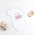 Baby onesie with floral design and text, accompanied by baby shoes and a teething ring on a light background