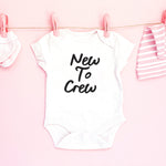 White baby onesie with 'New To Crew' text on a pink background