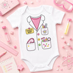 A white short-sleeve baby bodysuit with a cartoon scientist jacket graphic, featuring pockets with pens, a heart-shaped magnifying glass, and a yellow 'Scientist BABY' badge. The bodysuit is displayed in a flat-lay arrangement on a pink background with science-themed props like a ruler, notebook, and test tubes.