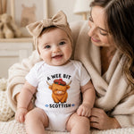 A baby wearing a white bodysuit with a Scottish highland cow graphic and the text 'A WEE BIT SCOTTISH' sits on a cozy cream knit blanket next to a smiling woman in a beige cardigan.