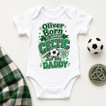 A white short-sleeve baby bodysuit laid flat on a light background, printed with green and gray text reading 'Oliver Born to support CELTIC Just like DADDY' around a soccer ball graphic. A small soccer ball, a Celtic coin, and a plaid fabric strip are placed around it as styling props.