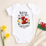 A white short-sleeve baby bodysuit laid flat on a light beige background, printed with a red dragon illustration and the text 'Kaira A Lil' Bit Welsh.' Yellow flowers and a small Welsh flag are placed around it as styling props.