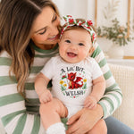 A smiling baby wearing a white bodysuit with a red and green fox graphic and the text 'A Lil' Bit Welsh' sits on a woman's lap. The woman wears a green and white striped long-sleeve shirt, and the baby also wears a matching red, green, and white floral bow headband.