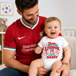 A man wearing a red Liverpool soccer jersey sits on a couch holding a baby wearing a white bodysuit with a red and green 'Born in Support Liverpool Just love DADDY' graphic.