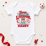A white short-sleeve baby bodysuit laid flat on a light beige background, printed with a red and teal graphic that reads 'Born to support Liverpool Just Like Daddy' with the Liverpool crest and soccer theme. Decorative red ribbon, star confetti, and a soccer ball are placed around the bodysuit.