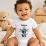 A smiling baby sitting on a white textured blanket, wearing a white short-sleeve bodysuit with a colorful graphic print that reads 'Siya Jean 100% CUTE' and features a baby bottle design with Australian and Nigerian flag colors. A teddy bear sits to the left, and a wooden toy chest is in the background.