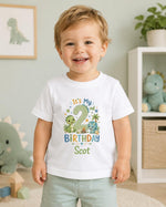 A smiling toddler boy wearing a white short-sleeve t-shirt with a colorful dinosaur-themed 'It's My 2nd Birthday' graphic, standing in a child's bedroom.