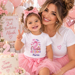 Woman with a smiling child wearing a personalised second birthday T-shirt and pink tutu beside a birthday cake.