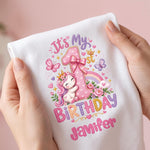 A white baby T-shirts held by two hands, featuring a colorful first birthday graphic with a pink unicorn, rainbow, butterflies, and the text 'It's My 1st Birthday Janifer'.
