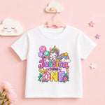 A white short-sleeve kids' t-shirt with a colorful cartoon unicorn and 'Jessica Turns One' text, displayed on a pink hanger against a pink background with decorative clouds and stars.