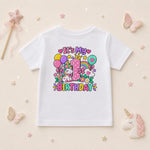 A white short-sleeve kids' t-shirt with a colorful 'It's My 1st Birthday' unicorn graphic, displayed on a light beige background with decorative pastel props including butterflies, a wand, and a unicorn plush.