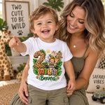 A toddler wearing a white graphic t-shirt with a safari-themed '2 Wild Beavers' print, sitting on a smiling woman in an olive green ribbed tank top in a cozy, decorated room.