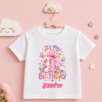 A white short-sleeve baby t-shirt with a colorful first birthday unicorn graphic, hanging on a pink hanger against a soft pink background with decorative cloud and star props.