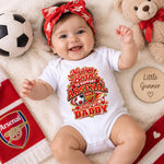 A baby lying on a cream blanket wearing a white short-sleeve bodysuit with a colorful Arsenal soccer graphic and a red bow headband. Surrounding props include a soccer ball, a teddy bear, a red Arsenal scarf, and a wooden name plaque.
