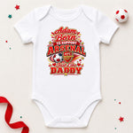 A white short-sleeve baby bodysuit laid flat on a light beige background, featuring a colorful graphic that reads 'Adam Born to Support Arsenal Just like Daddy' with the Arsenal FC crest and soccer ball motif. Decorative red ribbon, star confetti, and a small soccer ball are placed around the product.