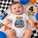 Smiling baby wearing a personalised 4 months old racing bodysuit beside toy cars and a wooden month marker.