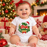 A baby sitting on a white shag rug in a cozy Christmas setting, wearing a white short-sleeve bodysuit with a festive graphic that reads 'My First Christmas 2024' in green, red, and gold. The baby is also wearing a red bow headband.