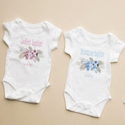 Two white baby onesies with floral designs and text on a beige background