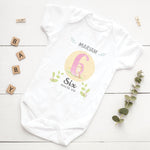 White baby onesie with milestone design on a wooden surface with decorative elements.