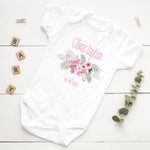 White baby onesie with floral design and text on a wooden surface with decorative elements.