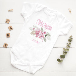 White baby onesie with floral design and text on a wooden surface with decorative elements.