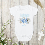 White baby onesie with floral design and text on a wooden surface with flowers and a cross.