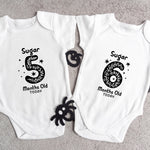 Two white baby onesies with black text and designs on a gray surface