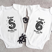 Two white baby onesies with black text and designs on a gray surface