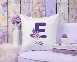 Decorative pillow with a purple letter 'E' and floral design on a floral-patterned wall background.