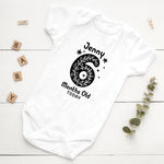 White baby onesie with black design and text on a white wooden surface with decorative elements.