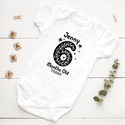 White baby onesie with black design and text on a white wooden surface with decorative elements.