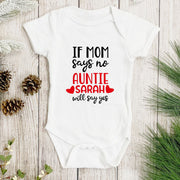 White baby onesie with text on a wooden surface with pine cones and greenery