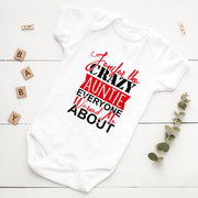 White baby onesie with red and black text on a white wooden surface with decorative elements.