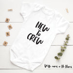 White baby onesie with 'NEW TO CREW' text on a white wooden surface with decorative elements.