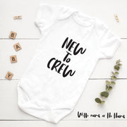 White baby onesie with 'NEW TO CREW' text on a white wooden surface with decorative elements.