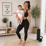 Woman holding a baby in a white bodysuit with a sample fitness logo, in a home workout space.