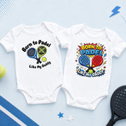 Two white padel baby bodysuits showing crossed racket artwork and a colourful “Born to padel like my Daddy” design.