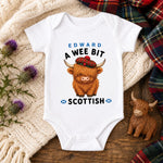 A white short-sleeve baby bodysuit laid flat on a wooden surface, printed with the text 'EDWARD A WEE BIT SCOTTISH' and a cartoon Highland cow wearing a red tartan hat. The bodysuit is styled with a cream knit blanket, purple thistle flowers, a small wooden Highland cow figurine, and a plaid scarf.