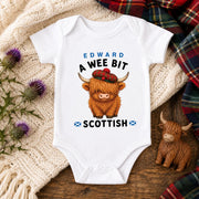 A white short-sleeve baby bodysuit laid flat on a wooden surface, printed with the text 'EDWARD A WEE BIT SCOTTISH' and a cartoon Highland cow wearing a red tartan hat. The bodysuit is styled with a cream knit blanket, purple thistle flowers, a small wooden Highland cow figurine, and a plaid scarf.