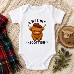 A white short-sleeve baby bodysuit laid flat on a cream knit blanket, printed with the phrase 'A WEE BIT SCOTTISH' and a cartoon Highland cow wearing a red tartan hat. A red plaid scarf, a small wooden Highland cow cutout, and thistle sprigs are arranged around the garment.
