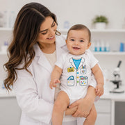 A baby wearing a white doctor-themed bodysuit with a medical coat and pocket graphic, held by a smiling woman in a white lab coat inside a bright, modern lab setting.