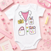A white short-sleeve baby bodysuit with a cartoon scientist jacket graphic, featuring pockets with pens, a heart-shaped magnifying glass, and a yellow 'Scientist BABY' badge. The bodysuit is displayed in a flat-lay arrangement on a pink background with science-themed props like a ruler, notebook, and test tubes.
