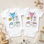 Two white baby bodysuits laid flat on a light beige surface, each printed with a cartoon scientist jacket design. One bodysuit has a blue inner shirt and yellow pocket labels, and the other has a pink inner shirt and yellow pocket labels.