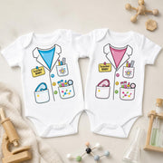 Two white baby bodysuits laid flat on a light beige surface, each printed with a cartoon scientist jacket design. One bodysuit has a blue inner shirt and yellow pocket labels, and the other has a pink inner shirt and yellow pocket labels.