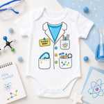 A white short-sleeve baby bodysuit laid flat on a light surface, printed with a cartoon scientist outfit featuring a blue lab coat, colorful pockets, and a yellow name tag reading 'Scientist BABY.' The bodysuit is styled with science-themed props including safety glasses, a beaker, molecular models, a notebook, and a card.