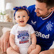 A baby wearing a white short-sleeve bodysuit with a colorful 'Jessica Born to Support Rangers Just like Daddy' graphic sits on a man wearing a blue Rangers FC jersey. The baby also wears a blue and red striped bow headband.