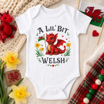 A white short-sleeve baby bodysuit laid flat on a beige fabric background, printed with a red dragon illustration and the text 'A LIL' BIT WELSH.' The bodysuit is surrounded by decorative props including yellow daffodils, a small red dragon toy, a crocheted textile, a Welsh flag, and a tartan fabric strip.