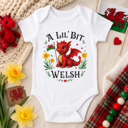 A white short-sleeve baby bodysuit laid flat on a beige fabric background, printed with a red dragon illustration and the text 'A LIL' BIT WELSH.' The bodysuit is surrounded by decorative props including yellow daffodils, a small red dragon toy, a crocheted textile, a Welsh flag, and a tartan fabric strip.