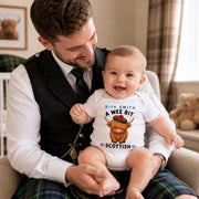 A baby wearing a white short-sleeve bodysuit with a Highland cow graphic and the text 'SIYA SMITH A WEE BIT SCOTTISH' sits on a man's lap. The man is wearing a black waistcoat, white shirt, tie, and tartan plaid trousers in a cozy living room setting.